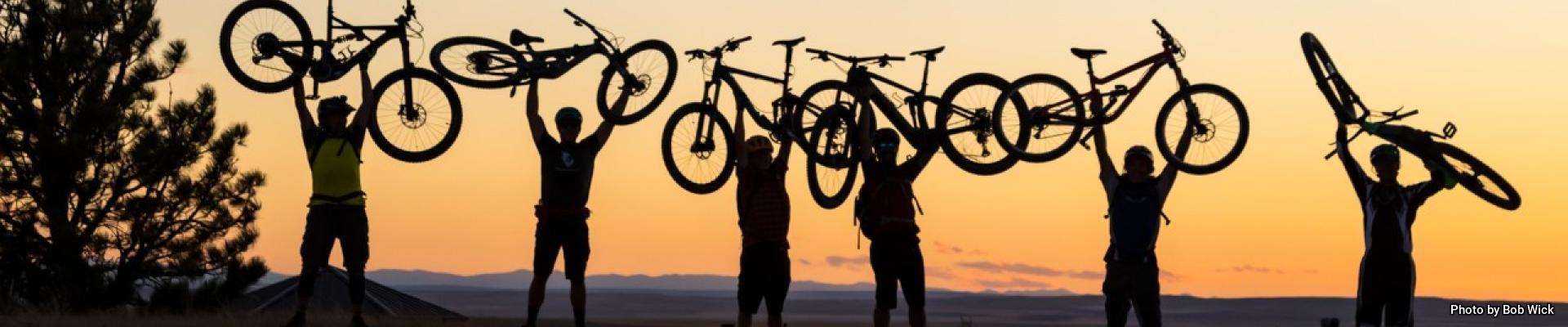 Silhouettes of a group of bicyclists as they hold up their electric bikes in front of a sunset in Montana's Acton Recreation Area | Photo by Bob Wick