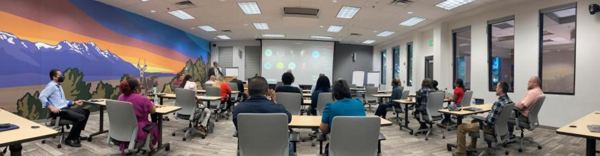 Group of people sitting in chairs behind tables listening to presentation in a National Training Center classroom with a large mural on the wall of a southwest landscape featuring a deer.